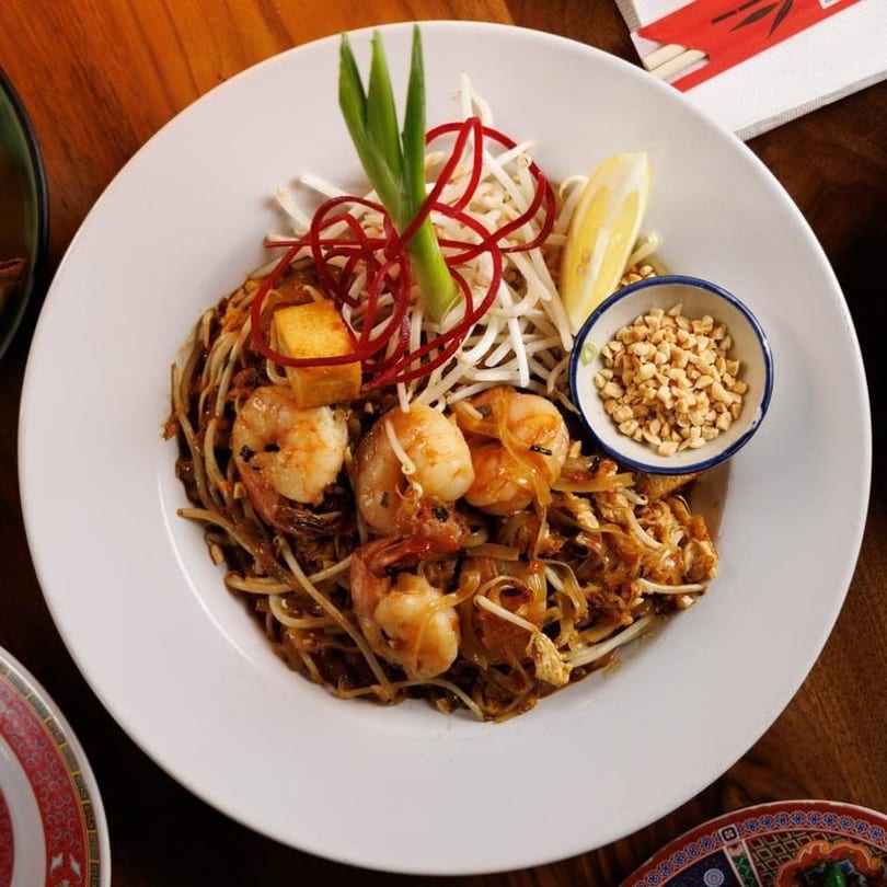 Shrimp pad thai with tofu, bean sprouts, red garnish, lemon, and peanuts.