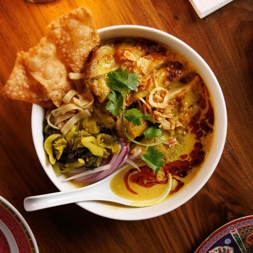 A bowl of Khao Soi with chicken, noodles, pickled greens, red onion, and crispy wontons.