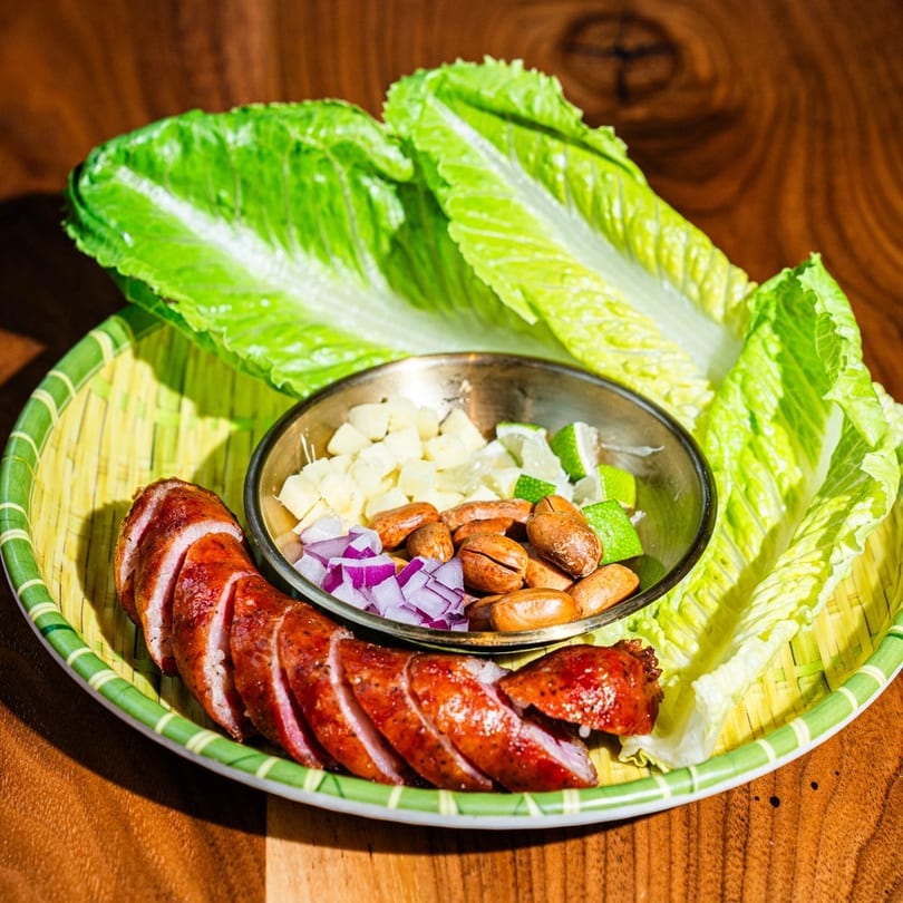 Sliced sausage, lettuce, and a bowl of peanuts, diced onions, and lime on a bamboo tray.