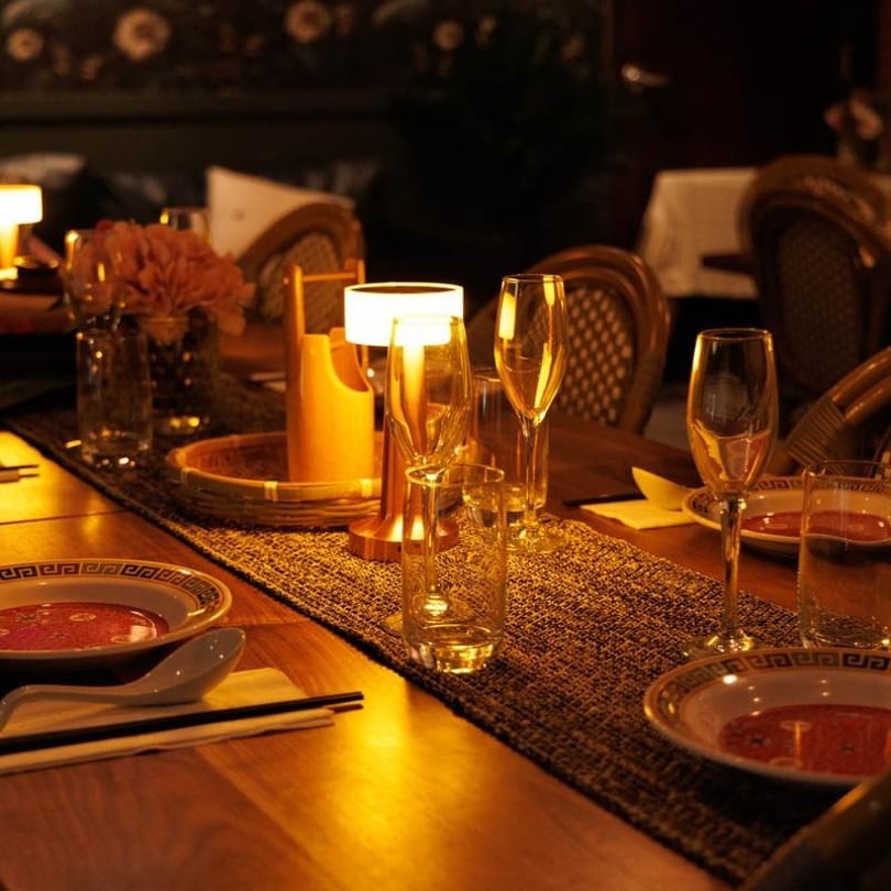 Elegantly set dining table with glowing lamps, wine glasses, and bowls of soup in a dimly lit restaurant.