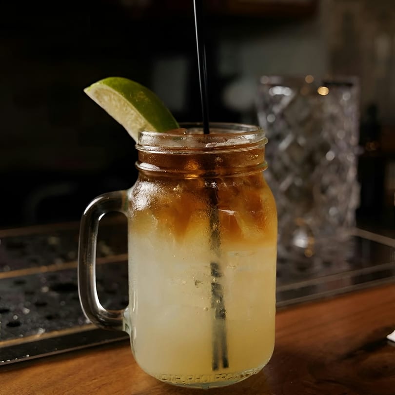A refreshing cocktail in a mason jar with a lime wedge and black straw on a wooden bar.