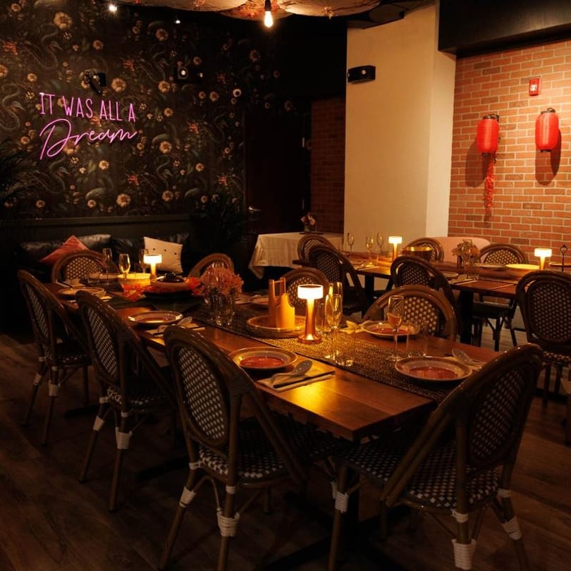 A restaurant dining room with a long table set with plates, glasses, and glowing lamps.
