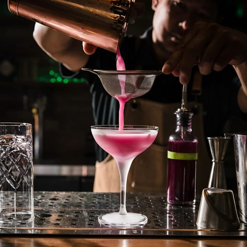 Bartender straining a vibrant pink cocktail from a copper shaker into a martini glass.