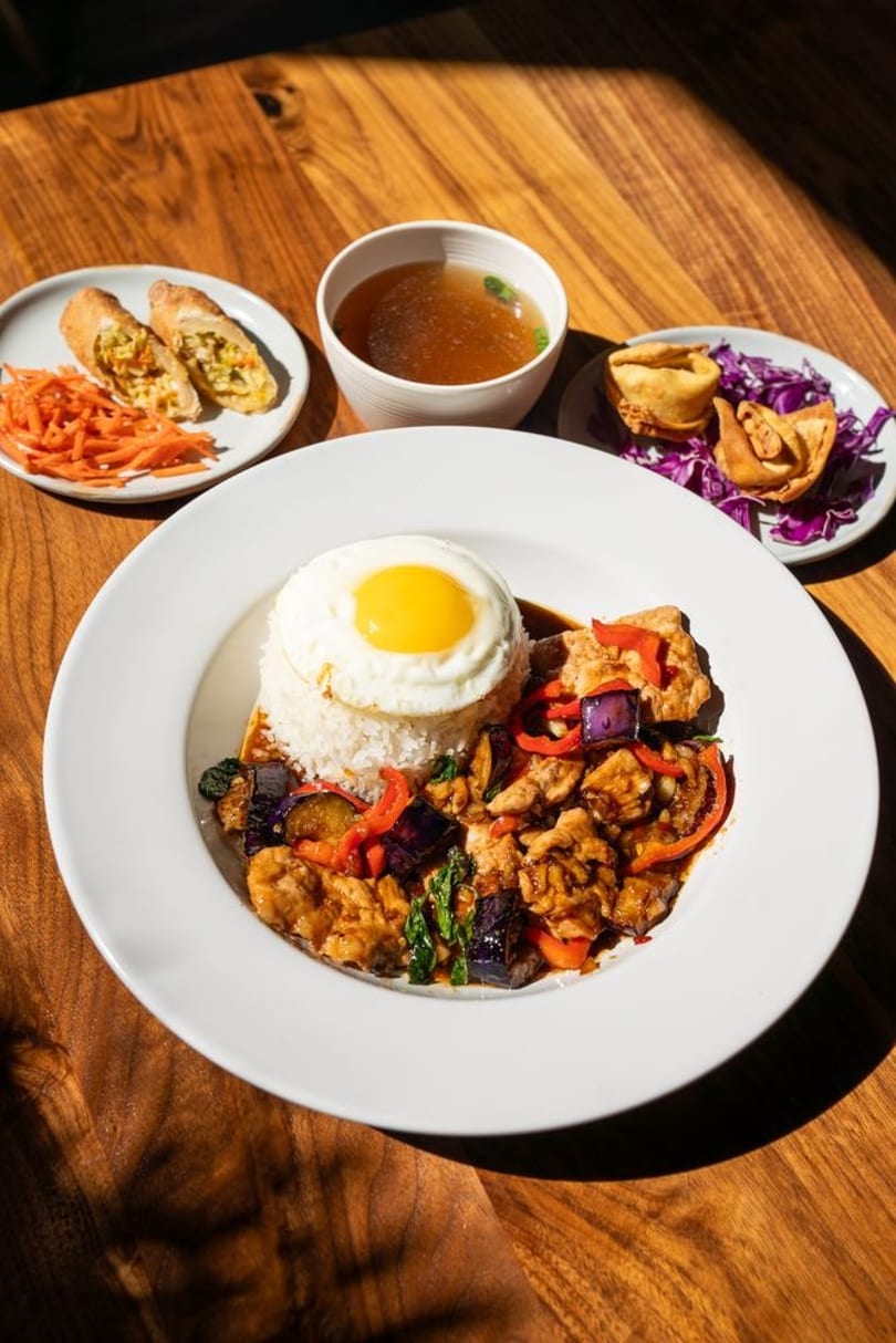 Thai basil chicken with fried egg, rice, soup, spring rolls, and fried wontons on a wooden table.
