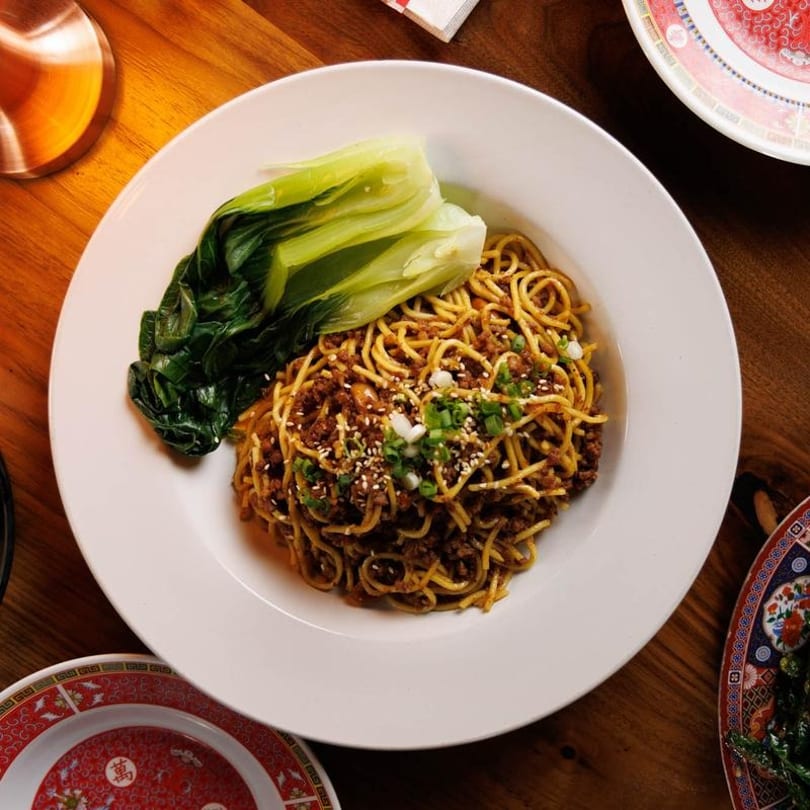 Dan Dan noodles with minced pork, bok choy, sesame seeds, and green onions in a white bowl.