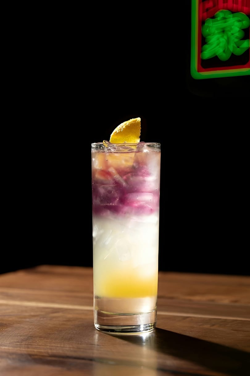 Layered cocktail with yellow, white, and purple liquids, garnished with a lemon slice on a wooden table.