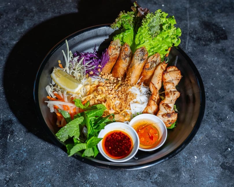 Vietnamese vermicelli bowl with spring rolls, grilled chicken, fresh vegetables, and dipping sauces.
